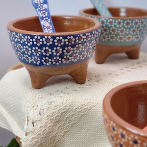 May include: Hand-painted ceramic bowls with matching spoons. The bowls are brown with a blue, white, and red dotted pattern. Each bowl has three small legs. The spoons have a similar design. The bowls are on a cream-coloured, woven surface.
