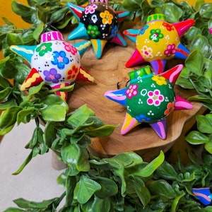 May include: Colorful star-shaped ornaments with floral designs. Each ornament features a different color scheme, including yellow, green, white, and blue, with vibrant accents. The ornaments are arranged on a wooden surface, surrounded by green foliage.