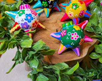 May include: Colorful star-shaped ornaments with floral designs. Each ornament features a different color scheme, including yellow, green, white, and blue, with vibrant accents. The ornaments are arranged on a wooden surface, surrounded by green foliage.