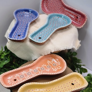 May include: A collection of colorful ceramic spoon rests. The spoon rests are in shades of blue, teal, coral, and yellow, each decorated with a floral pattern. The edges are trimmed with a white dotted design. A hole is at the end of each rest.
