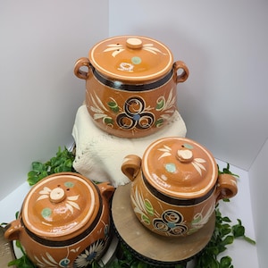 May include: Three ceramic cooking pots with lids. The pots are a warm orange-brown color with black and white accents. Each pot has a decorative floral design in blue, green, and white. The pots have handles and are displayed on a white surface and a wooden stand.