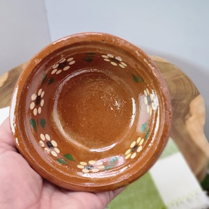 May include: A small, round, terracotta-colored bowl with a floral pattern. The rim is decorated with white and yellow daisy-like flowers and green leaves. The bowl has a glossy finish and is likely handmade.