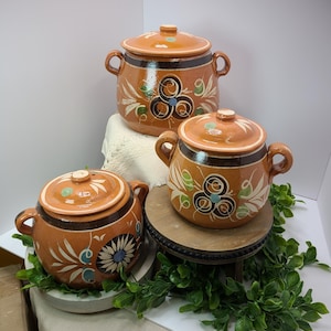May include: Three terracotta-colored ceramic pots with lids, adorned with floral and geometric designs in white, blue, and green. Each pot has two handles. The pots are arranged on a white surface with greenery.