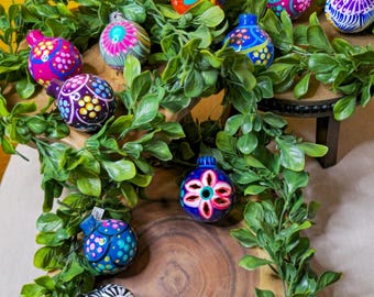 May include: A collection of colorful, hand-painted ceramic ornaments with intricate designs. The ornaments are arranged on wooden platforms and surrounded by green foliage.