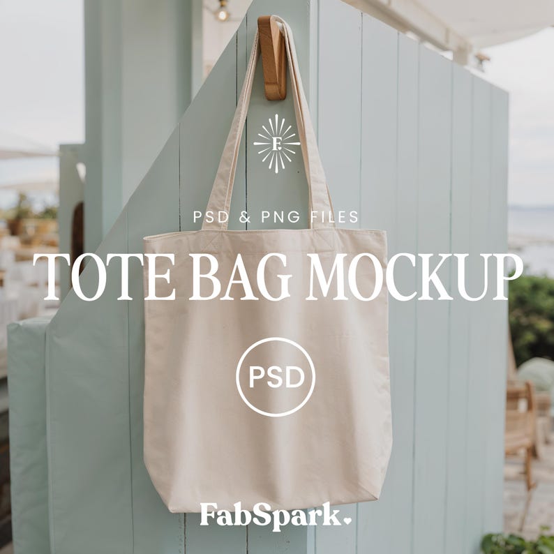 Pode incluir: Maquete de sacola tote bege pendurada em um gancho de madeira contra uma parede azul clara. A sacola tem um design simples com o texto "TOTE BAG MOCKUP" e "PSD & PNG FILES" em branco. O logotipo FabSpark est&aacute; na parte inferior.