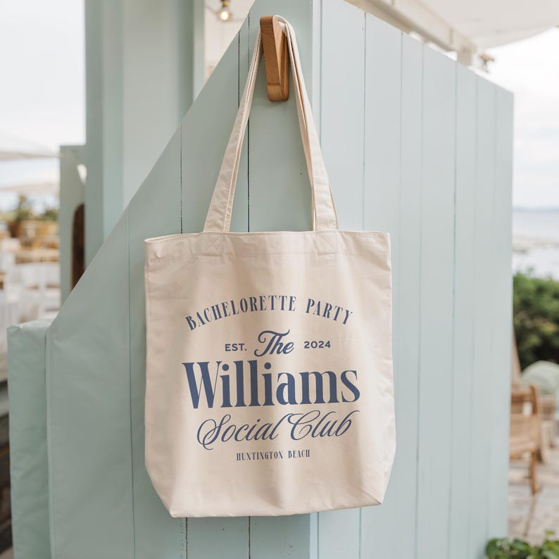 Pode incluir: Bolsa tote creme com texto azul marinho: "Bachelorette Party Est. The 2024 Williams Social Club Huntington Beach." A bolsa est&aacute; pendurada em um gancho de madeira em uma parede azul clara.