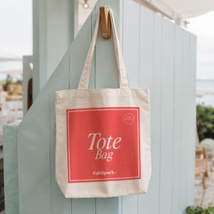 Pode incluir: Uma sacola tote creme com um design quadrado vermelho no centro, com as palavras "Tote Bag" em escrita branca e o logotipo FabSpark. A sacola est&aacute; pendurada em um gancho de madeira contra uma parede azul clara.