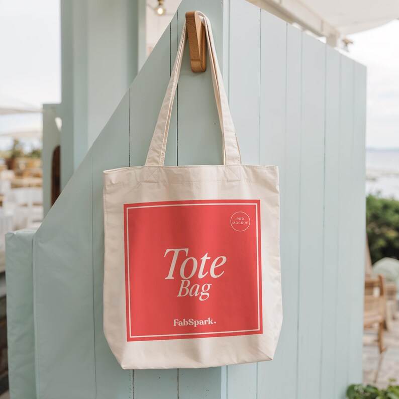 Pode incluir: Uma sacola tote creme com um design quadrado vermelho no centro, com as palavras "Tote Bag" em escrita branca e o logotipo FabSpark. A sacola est&aacute; pendurada em um gancho de madeira contra uma parede azul clara.