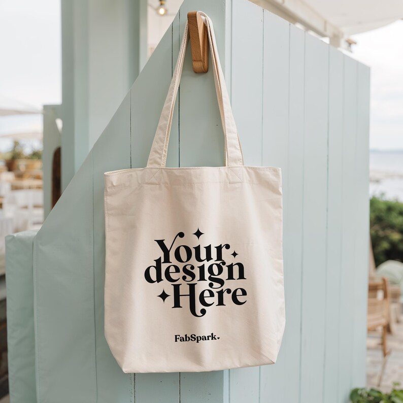 Pode incluir: Saco tote creme com texto preto que diz "Your design Here" e "FabSpark." O saco est&aacute; pendurado em um gancho de madeira em uma parede de madeira azul clara. O saco tem al&ccedil;as longas.
