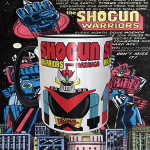 May include: White ceramic mug with a black handle. The mug features a colorful illustration of a robot from the "Shogun Warriors" comic book series. The text "Shogun Warriors Great Mazinga" is printed on the mug.