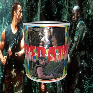 May include: A white ceramic mug with a black rim features a colorful image of characters from the movie Predator. The mug has the text "Predator The Hunt Will Begin" printed on it.