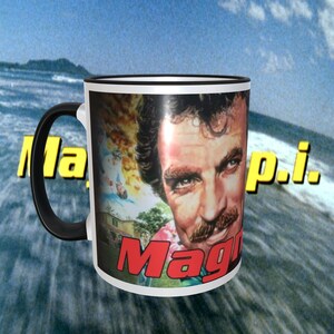 May include: White ceramic mug with a black handle and rim. The mug features a colorful image of a man with a mustache, and the words "Magnum P.I." in yellow and red lettering. The background is a blurred image of a beach scene.