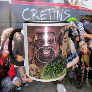 May include: White ceramic mug with a black rim and handle. The mug features a colorful graphic of a person with a large nose ring and the text "Class of Nuke High".