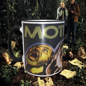 May include: A black and white ceramic coffee mug with a graphic of a man's face and the word "MOT" in yellow. The mug is sitting on a brown surface with a green plant in the background.