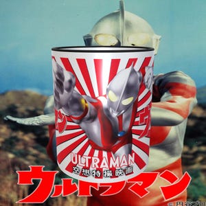 May include: A white ceramic mug with a red and white sunburst design. The mug features a graphic of Ultraman, a Japanese superhero, with the text "ULTRAMAN" and "宇宙特撮映画" in Japanese.