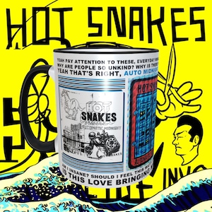 May include: A white ceramic mug with a black handle. The mug features a silver design with the text "Hot Snakes" and a phone graphic. The mug is surrounded by a yellow background with the text "Hot Snakes" in black.