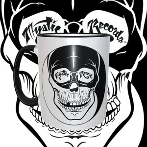 May include: White ceramic mug with a black rim and handle. The mug features a black and white skull design with the words "Mystic Records" in the eye sockets.