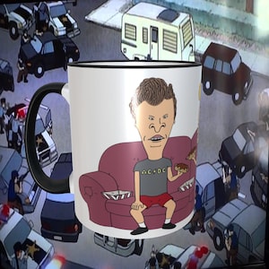 May include: A white ceramic mug with a black handle features a cartoon illustration of a character sitting on a pink couch. The character is wearing a grey t-shirt with the text "AC/DC" printed on it. The character is holding a hot dog in their right hand.