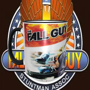 May include: White ceramic mug with a black rim and handle. The mug features a graphic of a helicopter and a car with the text "The Fall Guy" in red and yellow letters. The text "Stuntman Assoc." is printed in black letters on the bottom of the mug.