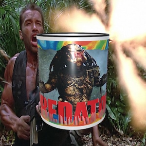 May include: A white ceramic mug with a black rim and handle. The mug features a colorful image of the Predator from the movie Predator, with the word "PREDATOR" printed in red letters below the image.