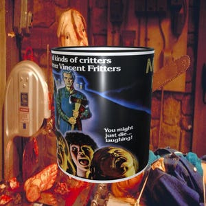 May include: A black and white mug with a movie poster design featuring a man with an axe and the text "all kinds of critters" and "mer Vincent Fritters". The poster also includes the text "You might just die... laughing!" and the letter "M".