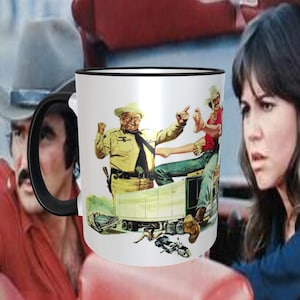 May include: A white ceramic mug with a black rim and handle. The mug features a colorful illustration of two men in a truck. The men are wearing cowboy hats and one is holding a gun. The text "Smokey and the Bandit" is printed on the mug.