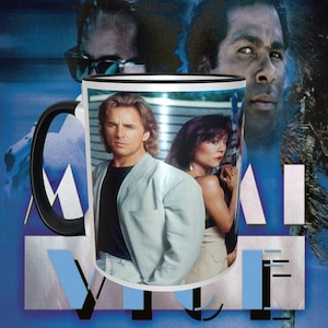 May include: White ceramic mug with a black handle. The mug features a photo of two people from the TV show Miami Vice. The mug has a black rim.