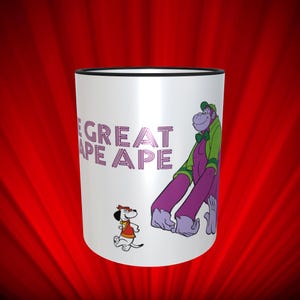 May include: White ceramic mug with a black rim. The mug features a cartoon image of The Great Ape Ape from the 1970s Hanna-Barbera cartoon. The ape is purple and wearing a green jacket and a green bow tie. A small dog with a red and yellow costume is running to the left of the ape. The text "The Great Ape Ape" is printed in pink on the mug.