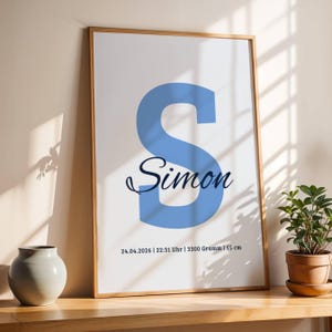 Könnte beinhalten: Gerahmter Kunstdruck mit einem großen, hellblauen Buchstaben "S" und dem Namen "Simon" in Schreibschrift. Darunter Datum, Uhrzeit, Gewicht (3300 Gramm) und Größe (55 cm). Der Rahmen ist hellbraun.