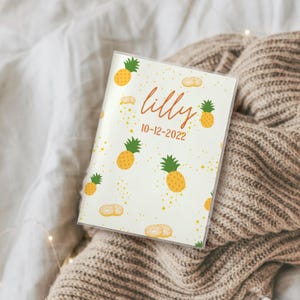 Könnte beinhalten: Ein personalisiertes Notizbuch mit Ananasmuster. Das cremefarbene Cover zeigt gelbe Ananas mit grünen Spitzen, den Namen "lilly" und das Datum "10-12-2022" in orangefarbener Schrift. Das Notizbuch liegt auf einer beigen Strickdecke.