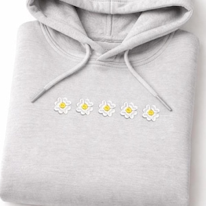 Può includere: Una felpa con cappuccio grigio chiaro con cappuccio con coulisse. Cinque margherite bianche ricamate con centri gialli sono allineate orizzontalmente sul petto. La felpa con cappuccio è piegata ordinatamente, mostrando il design.