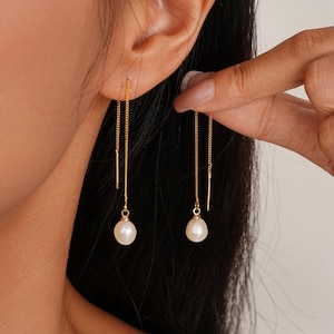 Könnte beinhalten: Zarte goldene Ohrringe mit einer kleinen, runden Perle am Ende jeder Kette. Die Ohrringe werden in einer Nahaufnahme gezeigt, die ihr elegantes Design hervorhebt. Die Ohrringe werden von einer Person hochgehalten.