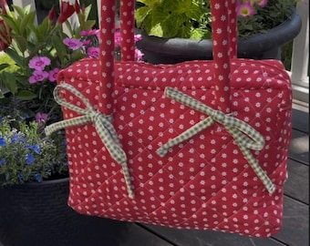 May include: Red quilted tote bag with white floral pattern and green gingham bows. The bag has matching red handles and is held up in front of a garden setting with flowers in pots.
