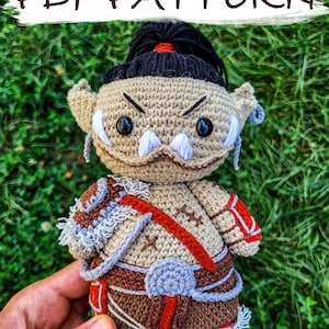 May include: Crocheted amigurumi toy of an Orc warrior with a brown, red, and gray outfit. The toy has white fangs and black eyes. The toy is being held in a hand against a green grass background. The text "PDF PATTERN" is at the top of the image.