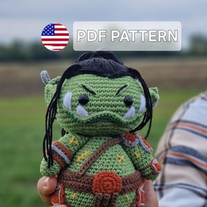 May include: A crocheted green orc doll with black hair and a brown strap across its chest. The doll has orange and brown accents and is held in a person's hand. The text "PDF PATTERN" is displayed above the doll.