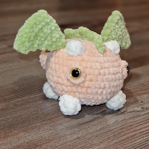 May include: A handmade crochet plush toy in the shape of a creature. The toy is peach-colored with white accents, green wings, and black and gold eyes. The plush has four white feet and white spots.
