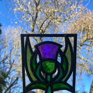 May include: A stained glass window panel depicting a purple thistle with green leaves. The thistle is a traditional symbol of Scotland.
