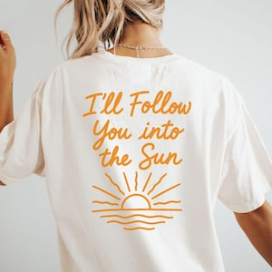 May include: Ivory-colored t-shirt with the text "I'll Follow You into the Sun" in orange script. Below the text is an orange graphic of a sun setting over the water. The word "IVORY" is printed at the bottom of the shirt.