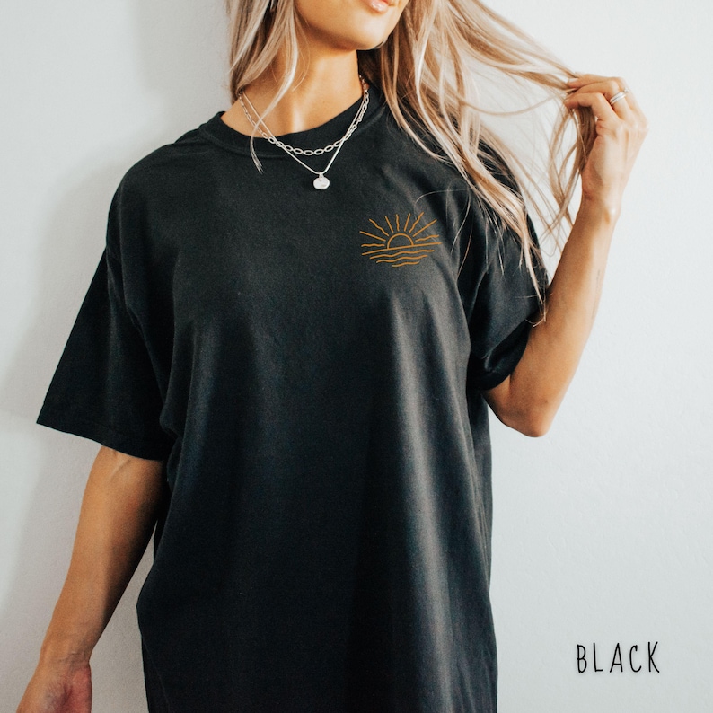 May include: Black t-shirt with a small embroidered design of a sun and waves in a golden-brown color on the upper left chest. The shirt has short sleeves and a relaxed fit. The word "BLACK" is printed in white on the lower right.