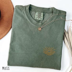 May include: A moss green t-shirt with a small embroidered sunset design in orange. The shirt has a crew neck and a small tag on the collar. A gold necklace with a pendant rests on the shirt. The word "MOSS" is at the bottom left.
