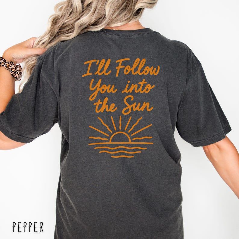May include: Dark grey t-shirt with the text "I'll Follow You into the Sun" in orange script. Below the text is an orange graphic of a sun setting over the ocean. The word "PEPPER" is printed in white at the bottom.