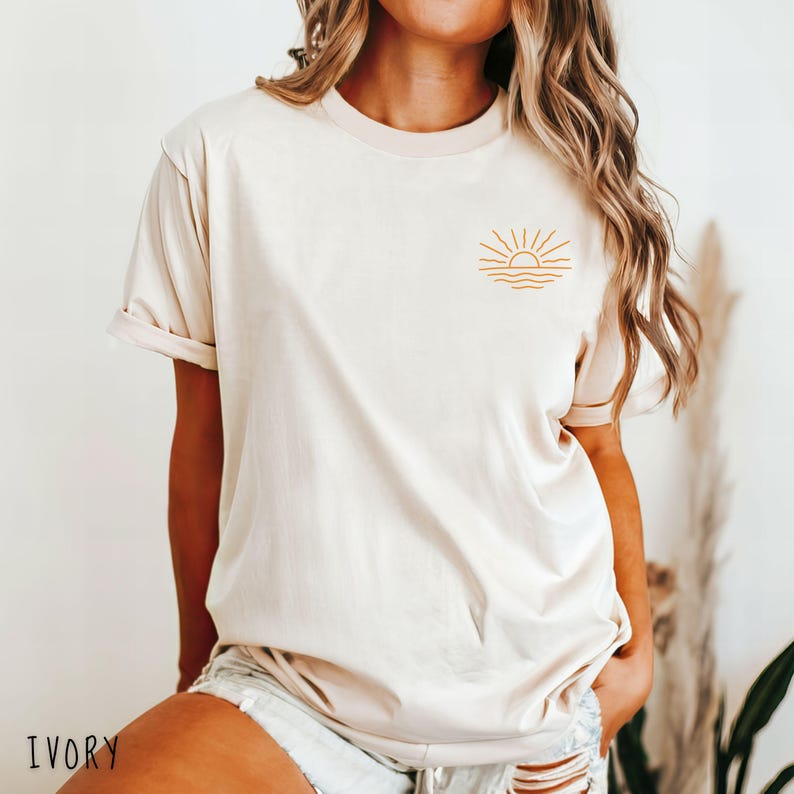 May include: Ivory-colored t-shirt featuring a minimalist sun and waves design in a golden hue. The short-sleeved shirt has a crew neck and a relaxed fit. The word "IVORY" is printed at the bottom of the image.