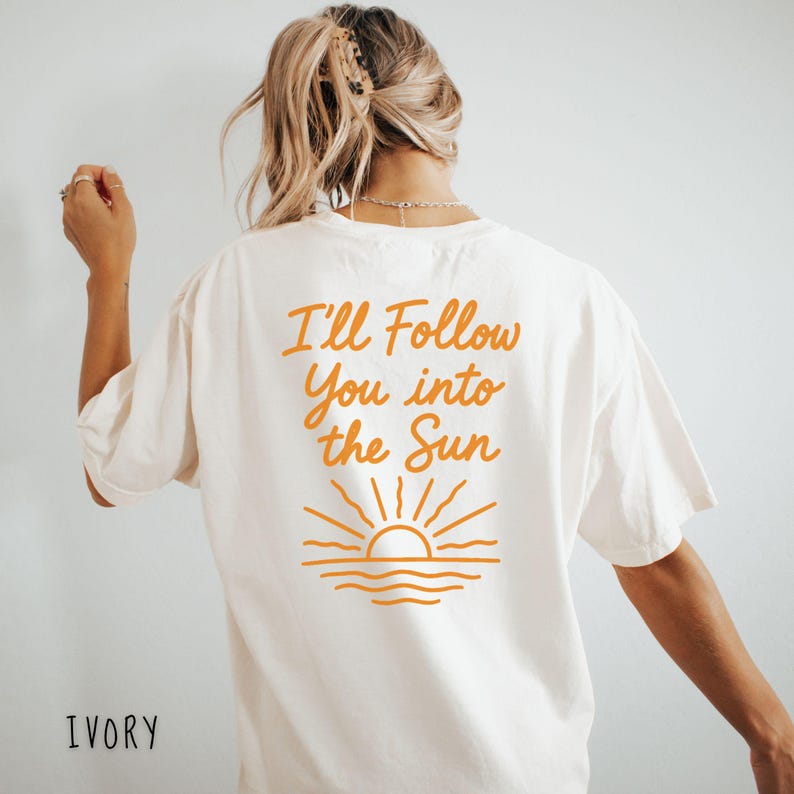 May include: Ivory-colored t-shirt with the text "I'll Follow You into the Sun" in orange script. Below the text is an orange graphic of a sun setting over the water. The word "IVORY" is printed at the bottom of the shirt.
