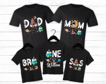 May include: Set of four black t-shirts with a space theme. The shirts feature a rocket, astronaut, planets, and stars. The text on the shirts reads "Dad", "Mom", "One William", and "Sis".