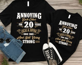 May include: Two black t-shirts with the text "ANNOYING Each Other FOR 20 YEARS LUCAS & SOPHIA SINCE 2003 And Still Going STRONG". One shirt is short-sleeved, the other is long-sleeved. A pair of blue jeans and black sneakers are also in the image.