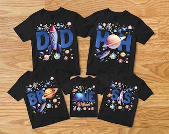 May include: Black t-shirts with space-themed designs. The shirts feature the words "DAD", "MOM", "BRO", "ONE", and "SIS" in blue, along with rockets, planets, and stars. The "ONE" shirt has the name "William".