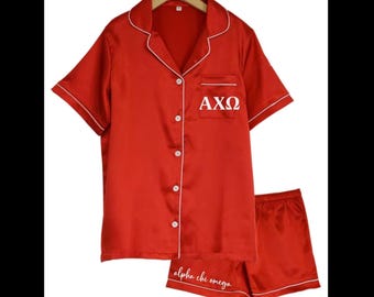 May include: A red satin pajama set consisting of a short-sleeved button-down shirt with a notched collar and matching shorts. The shirt features white piping and the Greek letters AXΩ in white. The shorts include the text Alpha chi omega in white.