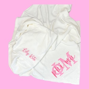 Op de afbeelding: Een witte, opgevouwen deken met de tekst "Lily Kate" in roze script op de capuchon. De Griekse letters "Phi Mu" worden ook in roze script op de deken weergegeven. De achtergrond is effen roze.