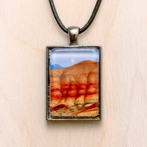 May include: A silver pendant necklace with a rectangular glass charm featuring a colorful landscape with red, orange, and yellow hills. The charm has a silver border and a black leather cord.