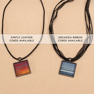 May include: Two necklaces with square glass pendants. The pendant on the left has a sunset-like design with orange, pink, and purple hues. The pendant on the right has a blue and white striped design. The necklace on the left is on a simple black leather cord. The necklace on the right is on a black organza ribbon cord. The text "SIMPLE LEATHER CORDS AVAILABLE" is above the left necklace. The text "ORGANZA RIBBON CORDS AVAILABLE" is above the right necklace.